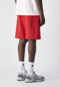 Czerwone spodenki sportowe z lekkiego materiału, w połączeniu z białymi skarpetami w prążki i szarymi trampkami. Prosty design, luźny krój, brak widocznych wzorów.