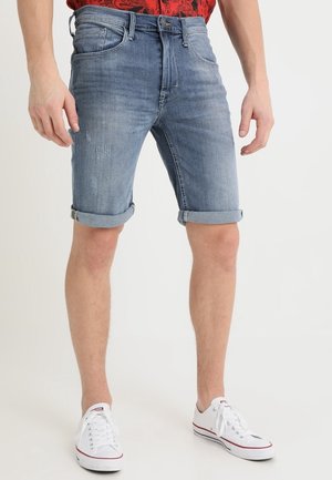 Homme portant un short en denim bleu retroussé et des baskets basses blanches, debout contre un fond clair uni.