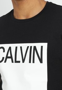 Zwarte katoenen t-shirt met een ronde halslijn, voorzien van een grote witte boxafbeelding met het woord "CALVIN" in vetgedrukte zwarte letters.