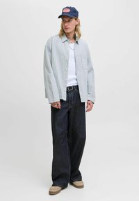 Karierter grün-weißer Hemd, weißes Tanktop, dunkle Jeans, helle Schuhe, dunkelblaues Cap mit Logo, stehende Pose.