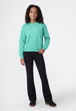 Jonge persoon met krullend haar, gekleed in een turquoise Champion-trui, zwarte broek en zwarte sneakers, staand tegen een witte achtergrond.