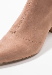 Bottine en suédine beige nude avec bout pointu, détails de coutures discrètes, et petit talon empilé, présentant une texture lisse.