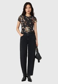 T-shirt in rete nera con fantasia floreale e maniche corte, abbinato a pantaloni in denim neri a vita alta. Modello tiene una borsa nera con una cintura.