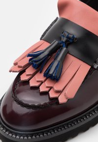 Mocassin en cuir bordeaux avec un empiècement à franges roses et des pampilles en cuir noir ornées de bords en daim bleu sur une semelle noire.