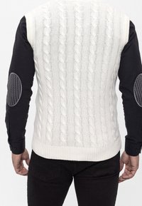 Gebreid crèmevest met kabelpatroon, aansluitend model, met zwarte lange mouwen en geruite elleboogstukken. Eindigt op middelhoge heuphoogte.