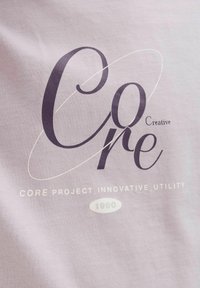 Šviesiai violetinė medvilninė marškinėliai su dideliu tamsiu tekstu, kuris sako "Core Creative" su spiralės grafika, o po jo yra "CORE PROJECT INNOVATIVE UTILITY".