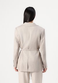 Hellbeige Blazer mit tailliertem Schnitt, Gürtel in der Taille, Reverskragen und Rückenschlitzdetails. Der Stoff hat eine subtile Textur.