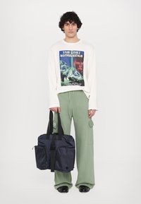 Navy blue fabric bag with black straps, two front pockets, and a central zip. Model wears a graphic long-sleeve shirt and green pants.