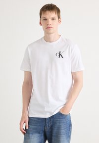 Bijela pamučna majica s okruglim izrezom i kratkim rukavima, s crnim "cK" logotipom na lijevoj strani prsnog koša. Casual kroj.