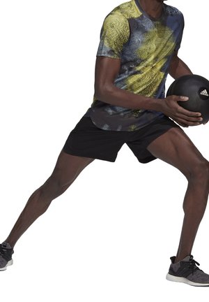 Sportshirt mit einem mehrfarbigen Muster in Gelb und Blau, kombiniert mit schwarzen Shorts und schwarzen Sportschuhen, hält einen schwarzen Medizinball.