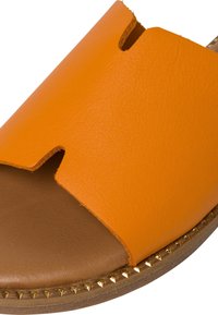Gros plan sur une sandale en cuir orange avec un design découpé, une semelle intérieure marron et des clous dorés en forme de pointes le long du bord de la semelle.