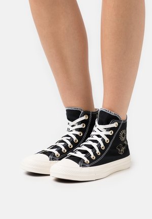 Wysokie trampki z czarnego płótna z białymi sznurowadłami i kontrastującymi przeszyciami; wyposażone w złote przelotki i dekoracyjne graficzne akcenty.