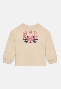 Μπεζ μπλούζα με μακριά μανίκια και στρογγυλή λαιμόκοψη. Φέρει το "GAP" με κόκκινα γράμματα και γραφικό "Hot Wheels Racing" με καρό σημαίες στο πίσω μέρος.