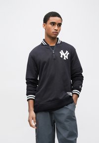 Fekete pulóver fehér logóval a bal mellkason, cipzáros gallérral, csíkos mandzsettákkal és bordázott szegéllyel. Puha anyagból készült.