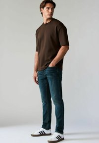 Barna, rövid ujjú póló sötét teal slim fit farmernadrággal és fekete csíkos, barna részletekkel ellátott fehér sportcipővel párosítva.