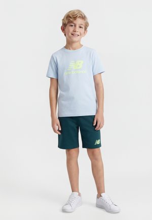 Ragazzo giovane in piedi, indossa una maglietta azzurro chiaro New Balance, pantaloncini verde scuro e scarpe da ginnastica bianche, sorride davanti a uno sfondo neutro.