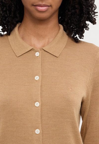 Beige neulejakku, jossa on kaulus, valkoiset napit ja hienovarainen logoyksityiskohta, joka esittelee sileää pintaa ja istuvaa muotoilua.