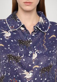 Chemise boutonnière bleu foncé avec des bordures blanches, ornée d'un motif fantasque d'étoiles, de vaches, de cygnes et d'éléments célestes sur le tissu.