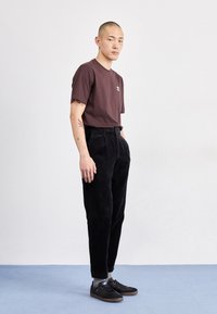 Vinröd bomullst-shirt, svarta manchestertygsbyxor och svarta sneakers. Personen står med händerna i fickorna på ett ljusblått golv.