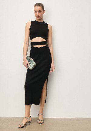 Moteris juodo be rankovių su šoniniu kirpimu suknelėje, rankoje laikanti mažą žalios gyvatės odos rašto delninukę, avinti derančius dirželiais tvirtinamus aukštakulnius.