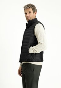 Gilet invernale nero con tasche con zip, dotato di colletto alto. Indossato sopra un maglione a coste beige chiaro. I pantaloni verde oliva completano l'outfit.