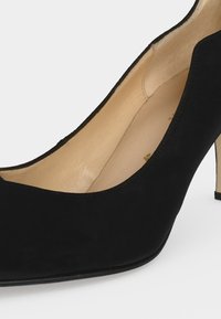 Escarpin en daim noir à talon haut avec bout pointu et doublure intérieure en cuir beige sur fond blanc.