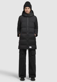 Geacă puffer neagră, cu fermoar în față, dotată cu glugă, baffluri mari și buzunare laterale. Purtată cu pantaloni cargo negri și o căciulă tricotată.