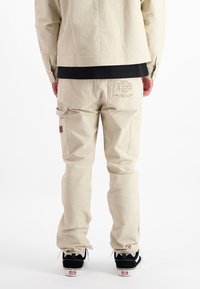 Beige cargobroek met zakken, voorzien van een gestructureerde stof, geborduurd logo op de achterkant, en zwarte schoenen met witte zolen.