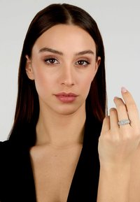 Anello d'argento con più file di piccole pietre scintillanti, indossato sulla mano destra. La modella ha lunghi capelli scuri e un trucco neutro.