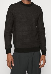 Maglione nero lavorato a maglia con un motivo testurizzato, scollatura rotonda e polsini e orlo a coste. Abbinato a pantaloni grigio scuro.