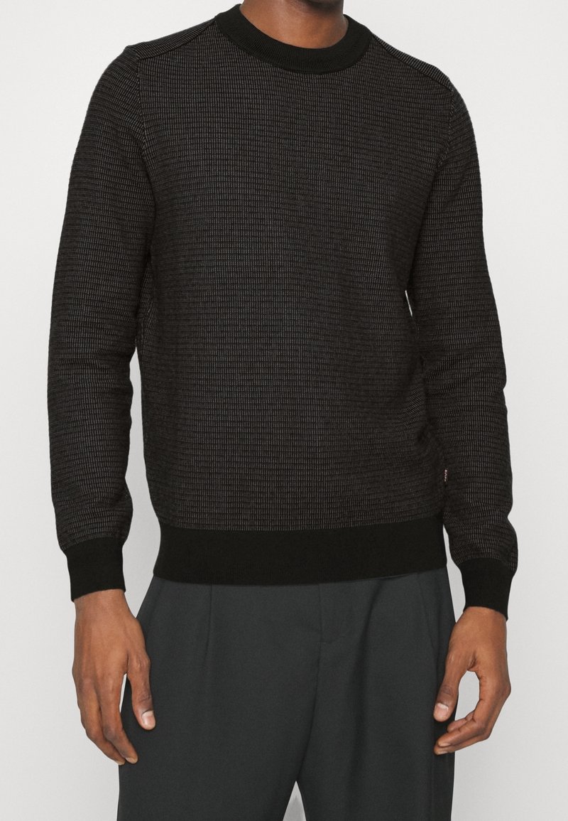 Maglione nero lavorato a maglia con un motivo testurizzato, scollatura rotonda e polsini e orlo a coste. Abbinato a pantaloni grigio scuro.
