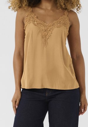 Kobieta ubrana w beżowy, bezrękawnik z koronkowymi detalami, dobrany do ciemnoniebieskich dżinsów, na białym tle.