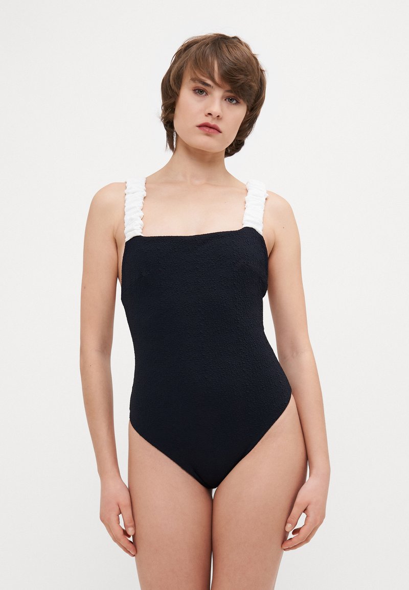 Black one-piece swimsuit with textured fabric, square neckline, and white ruffled straps; features high-cut leg openings.