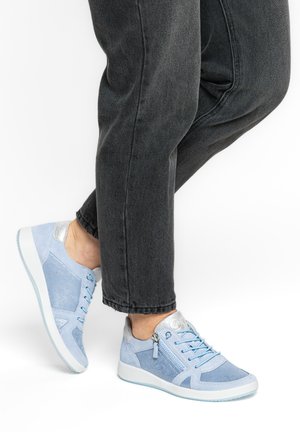 Hellblaue Sneaker mit Blumenmuster und silbernen Akzenten, getragen zu schwarzen Jeans, zeigen seitlichen Reißverschluss und Schnürsenkel auf weißem Hintergrund.