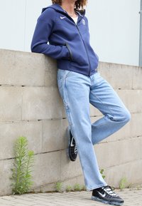 Un sweat à capuche zippé navy avec logo, un jean bleu clair ample, et des baskets noires. Le sweat comprend une capuche et des poches latérales, le jean a une coupe droite.