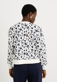 Mujer con cabello rizado corto, vistiendo una blusa blanca de mangas largas con manchas de leopardo en azul y negro, y pantalones oscuros, de espaldas.
