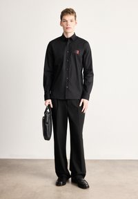 Μαύρο μακρυμάνικο πουκάμισο με κουμπιά, με μικρό κόκκινο λογότυπο, σε συνδυασμό με μαύρο παντελόνι και κρατώντας μαύρη δερμάτινη τσάντα.