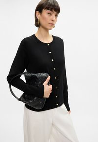 Cardigan nero a maglia con scollatura rotonda, chiusure con bottoni dorati e maniche lunghe. Il modello tiene una borsa intrecciata nera. Pantaloni bianchi visibili.