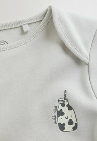 Šviesiai pilkas medvilninis kūdikių bodžiukas trumpomis rankovėmis, su atspausdintu piešiniu – besišypsančiu pieno buteliuku su šiaudeliu ir juodomis karvės dėmėmis.