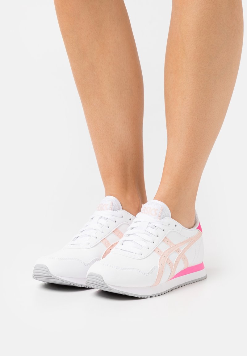 Vita och rosa Asics-sneakers på barfotade fötter mot en enkel vit bakgrund.