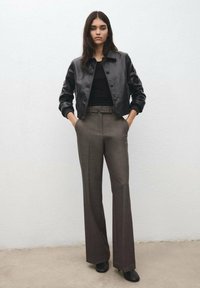 Kobieta stojąca z rękami w kieszeniach, ubrana w czarną skórzaną kurtkę, czarny top, szare spodnie z wysokim stanem i czarne buty, na tle białej ściany.