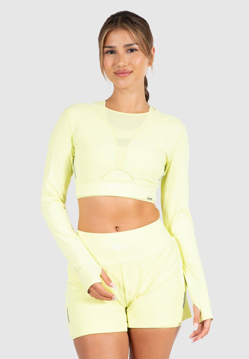 Neon-gelbes sportliches Crop Top mit langen Ärmeln und Netzdetails, kombiniert mit passenden Shorts, die seitliche Schlitze und Logodetails aufweisen.