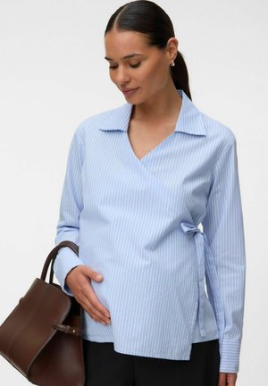 Donna incinta con una blusa a righe blu incrociata che tiene la pancia con una mano e una borsa marrone nell'altra.