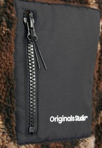 Brown and black textured fabric featuring a zippered pocket with a smooth zipper and white "Originals Studio" branding on the side.