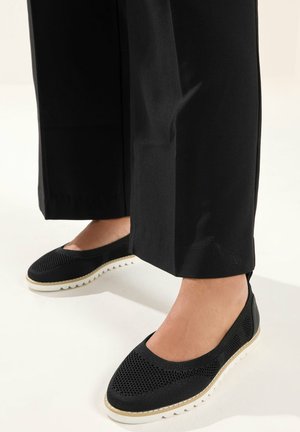 Piedi con indosso scarpe slip-on nere in maglia con suole bianche, abbinate a pantaloni neri a gamba dritta su uno sfondo chiaro.