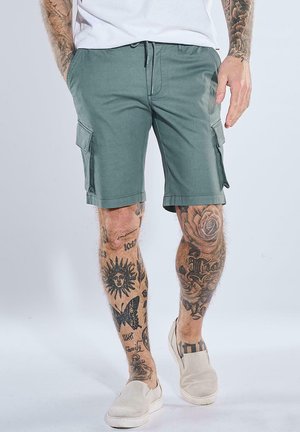Mann mit grünen Cargoshorts und weißen Slip-On-Schuhen mit tätowierten Beinen, steht vor einem schlichten Hintergrund.