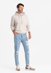 Beżowa bluza z kapturem z przednią kieszenią i sznurkiem, w połączeniu z jasnoniebieskimi dżinsami z przetarciami, które mają dziury. Uzupełniona o modne sneakersy.