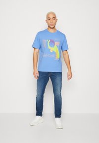 Camiseta de algodón azul con un diseño gráfico colorido que presenta formas abstractas y textos, combinada con unos jeans ajustados azules y zapatillas blancas.