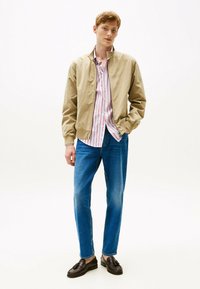 Chaqueta bomber caqui sobre una camisa de rayas rosas y blancas, combinada con vaqueros azules y mocasines marrón oscuro. Diseño simple y de corte recto.