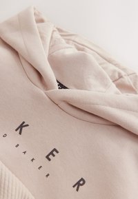 Šviesiai smėlio spalvos medvilnės mišinio gobtuvu megztinis, su tekstūruota plokštele ir ryškia juoda logotipų spauda priekyje. Lygus audinys, kasdieniškas dizainas.